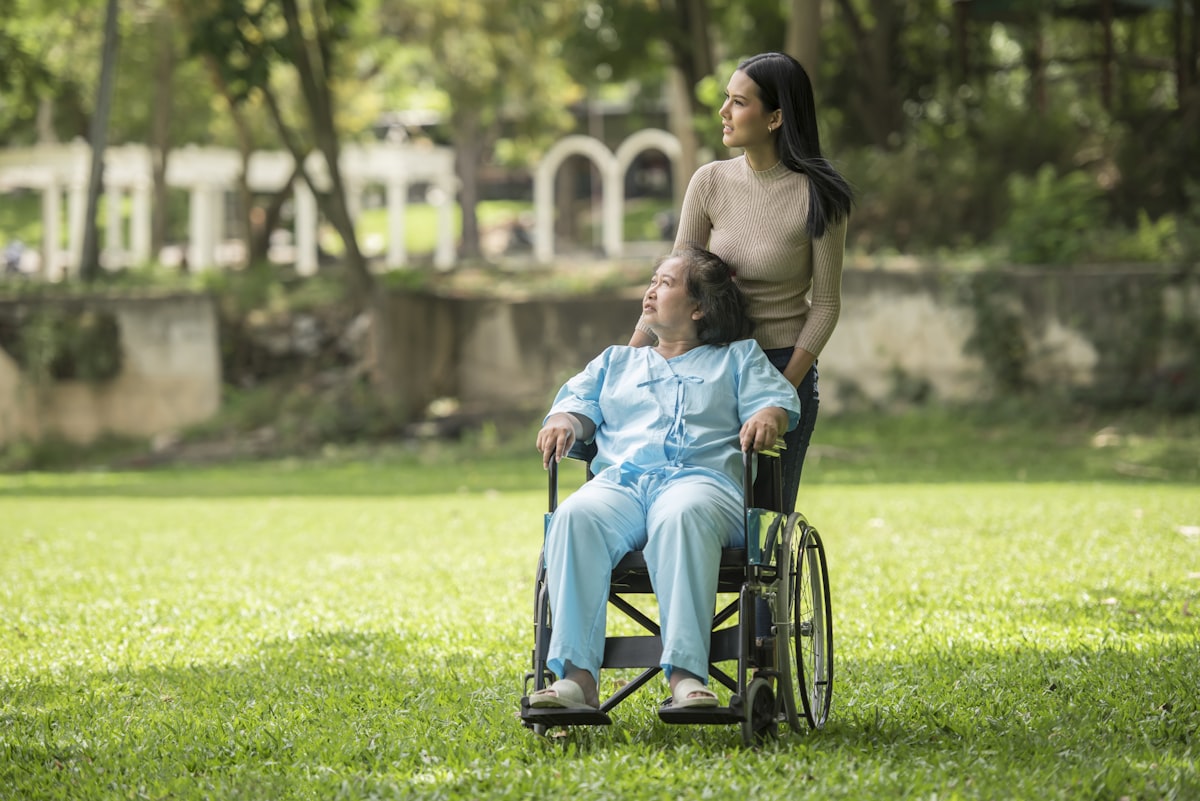 An Asian woman in a wheelchair in a sunlit park, with a younger Asian caretaker standing behind her